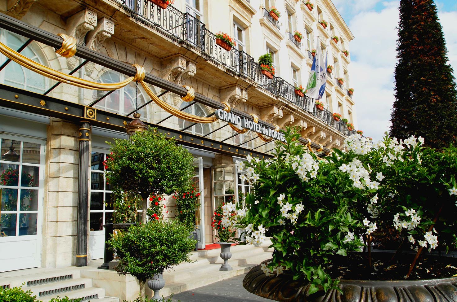 The exterior of the Intercontinental Bordeaux hotel with its white bricks and gold detailing on a bright day with greenery and white flowers in front.